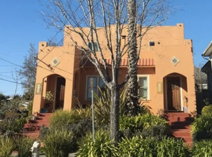 A bright orange house stands against the blue sky. Two front doors flank a palm tree and plants in front of it